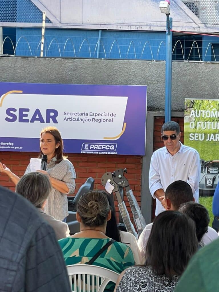 A prefeita Adriane Lopes discursa ao lado do subprefeito de Anhanduí, Elenilton Dutra, que veste camisa branca e óculos escuros, durante a entrega de equipamentos na SEAR.