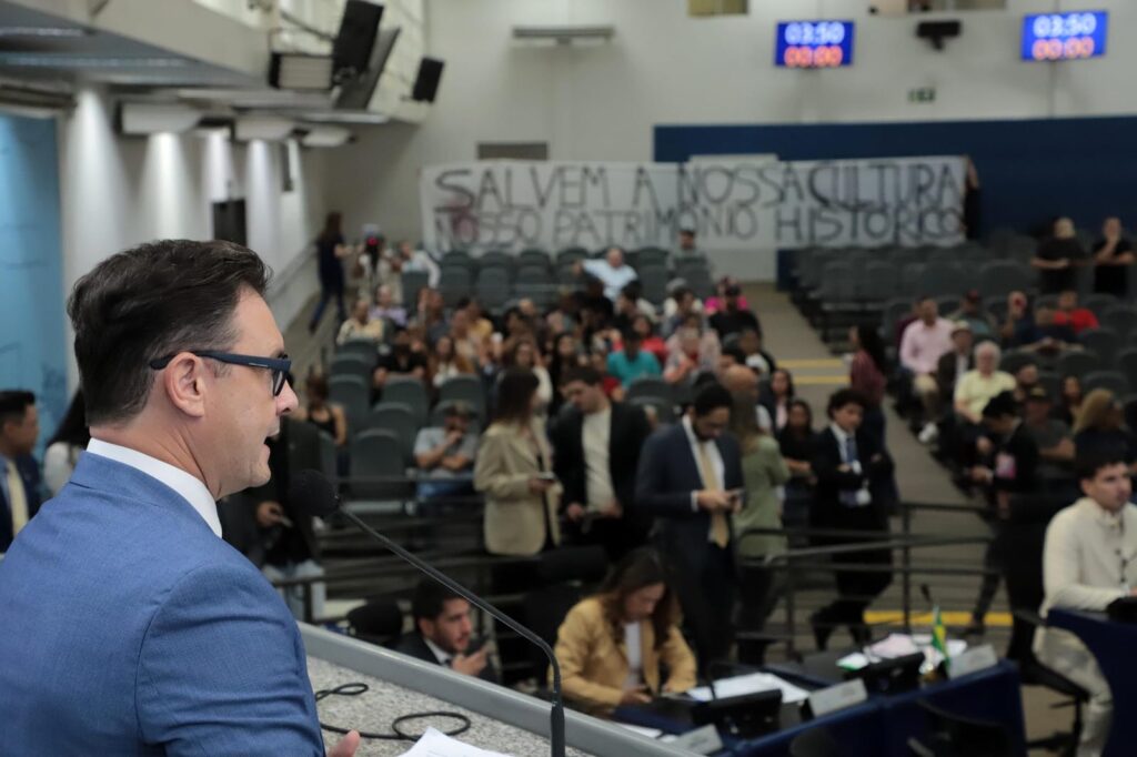 O vereador André Salineiro discursa ao microfone na tribuna da Câmara Municipal de Campo Grande durante a sessão plenária. Ao fundo, o público acompanha a votação e exibe uma faixa com os dizeres: 