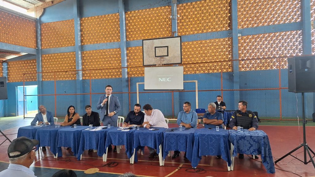 Audiência pública realizada no Ginásio da Escola Municipal Isauro Bento Nogueira, em Anhanduí, com comerciantes, moradores e representantes da comunidade reunidos em torno de uma mesa para discutir a preservação das atividades tradicionais. Um dos participantes fala ao microfone, enquanto autoridades e representantes da comunidade acompanham o encontro.