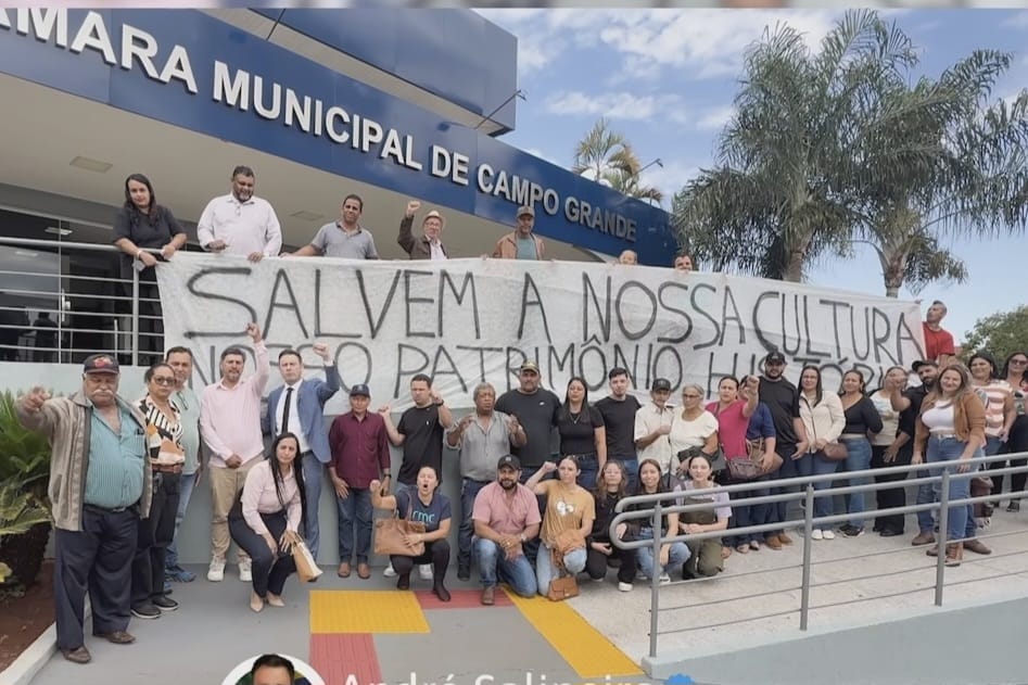 Grupo de moradores e participantes reunidos em frente à Câmara Municipal de Campo Grande, com Jezhel e Salineiro entre os presentes. O grupo segura uma faixa com a mensagem “Salvem a nossa cultura e o patrimônio”, em manifestação relacionada à preservação das atividades tradicionais de Anhanduí.